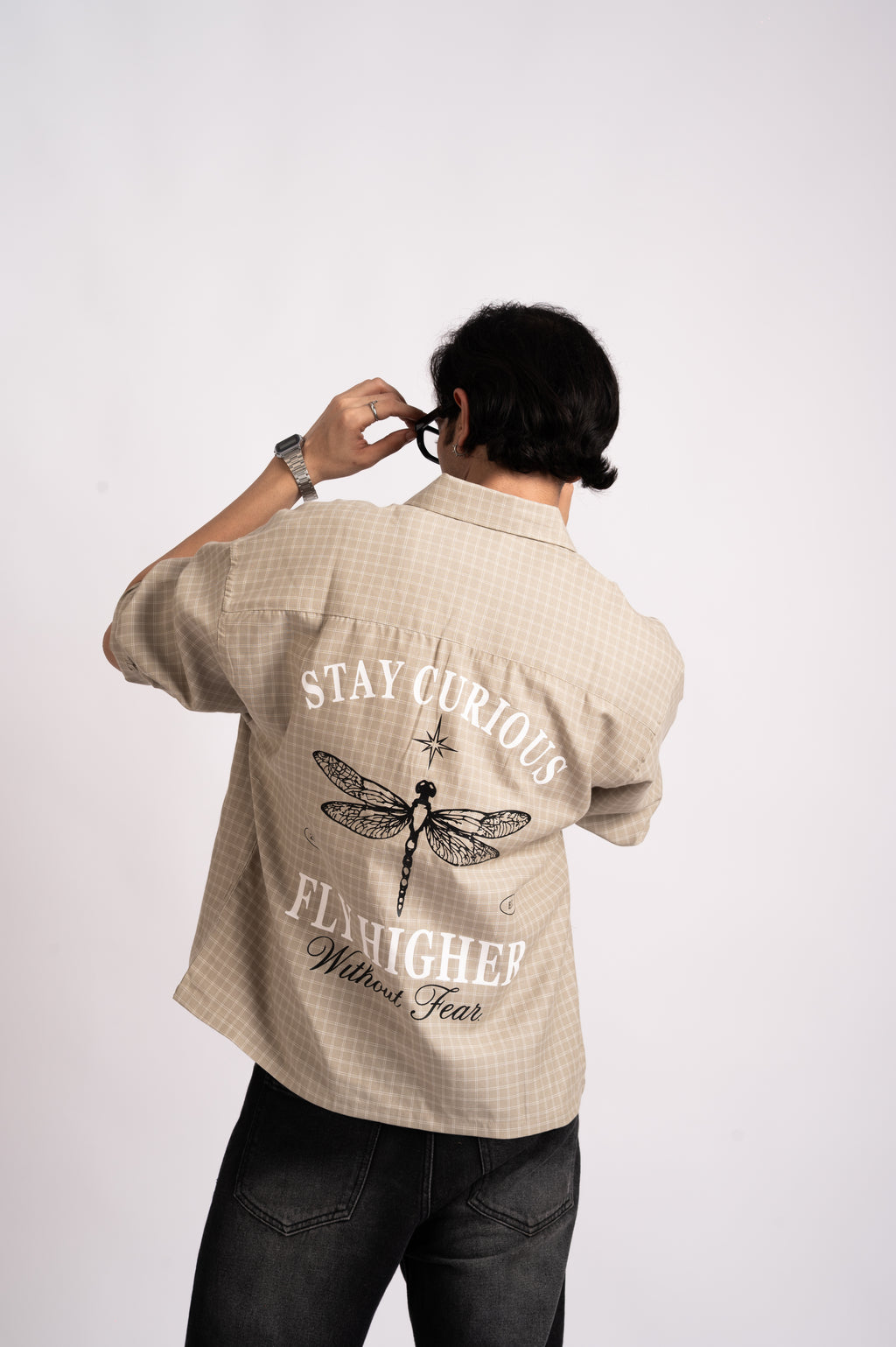 A person wearing a Nomad Check Boxy Shirt- Beige, a short-sleeved button-down with a dragonfly graphic and the text Stay Curious Fly Higher Without Fear printed on the back.