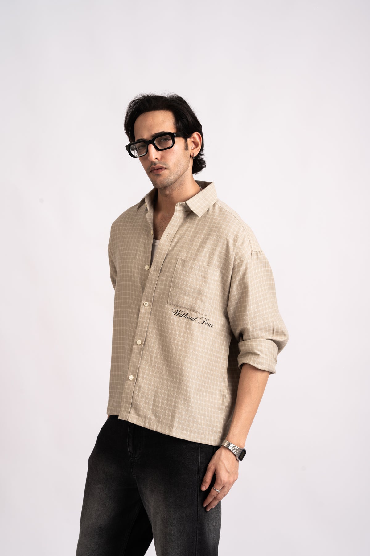 A man wearing a relaxed-fit beige Nomad Check Boxy Shirt with Without Fear embroidered on the pocket and black jeans.